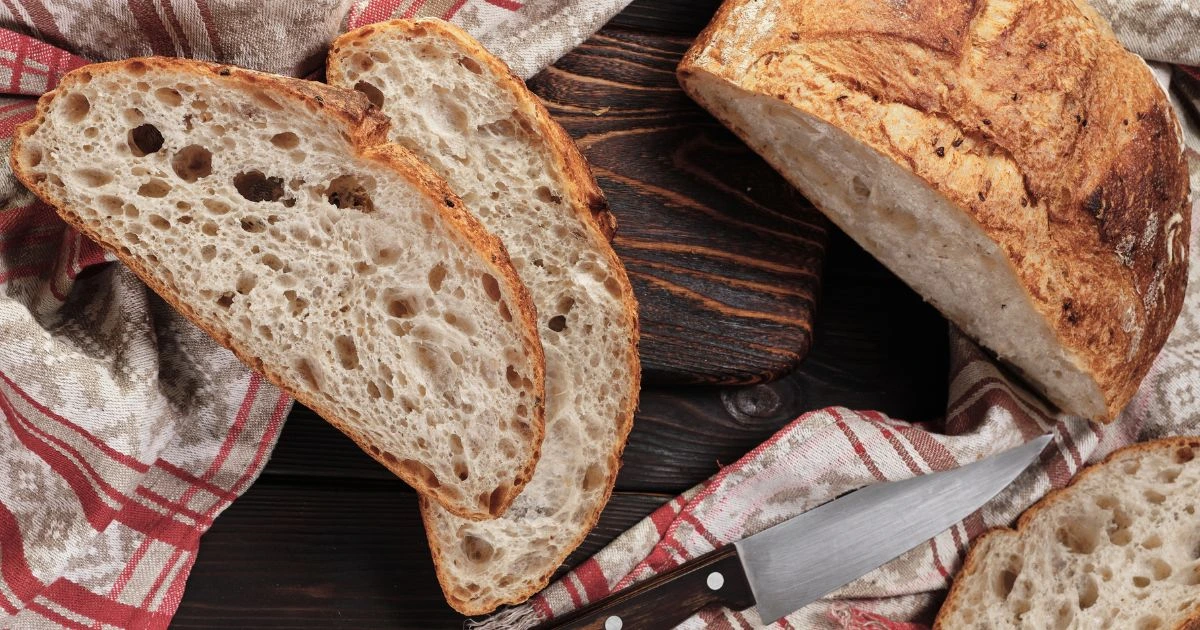 Sliced sourdough bread on a wooden surface with a knife and a red-checkered cloth.