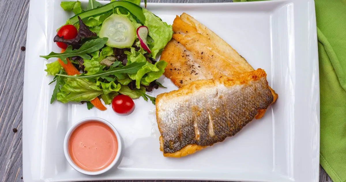 Grilled branzino fillet with crispy skin served alongside a fresh salad and dipping sauce.