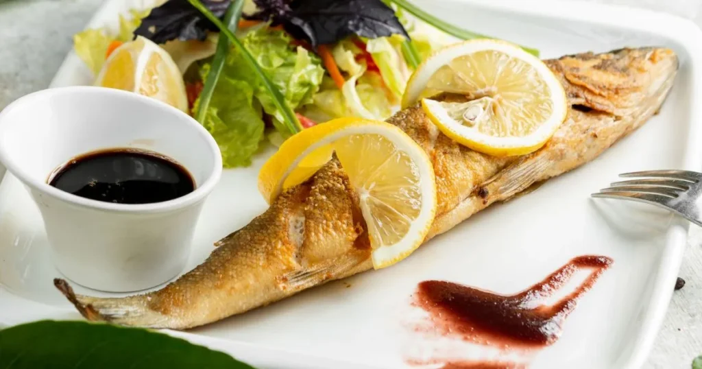 A perfectly cooked branzino served whole with lemon slices, a side of fresh salad, and dipping sauce on a white plate.