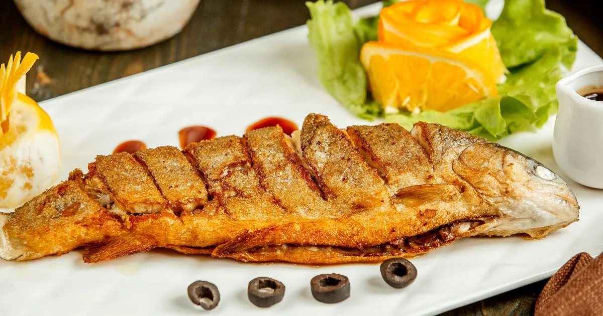 A perfectly cooked branzino fish, served whole with a golden crispy skin, accompanied by orange slices, fresh lettuce, and black olives on a white plate.