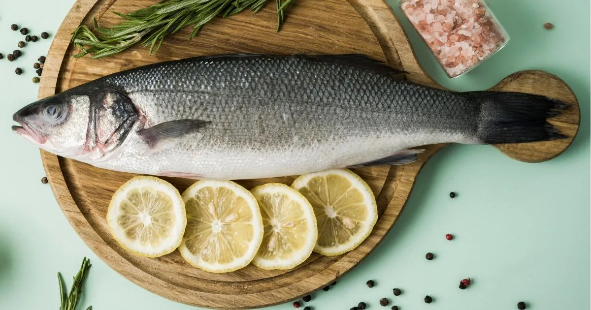 Fresh branzino fish on a wooden board with lemon slices, rosemary sprigs, pink Himalayan salt, and peppercorns.