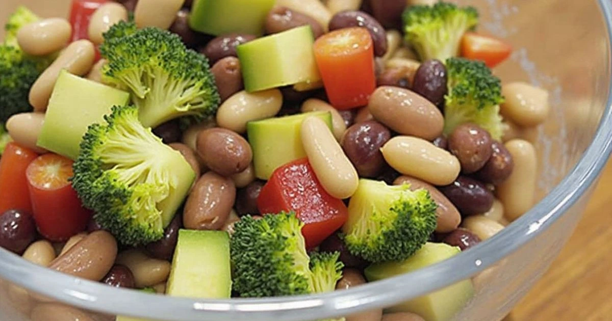 A vibrant salad featuring a mix of beans, broccoli florets, diced avocado, cherry tomatoes, and red bell peppers in a clear glass bowl.