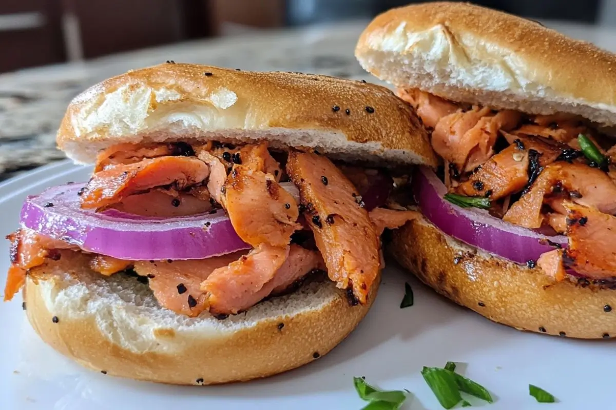 Close-up of two lox bagel sandwiches with smoked salmon, red onion slices, and fresh herbs on a poppy seed bagel.