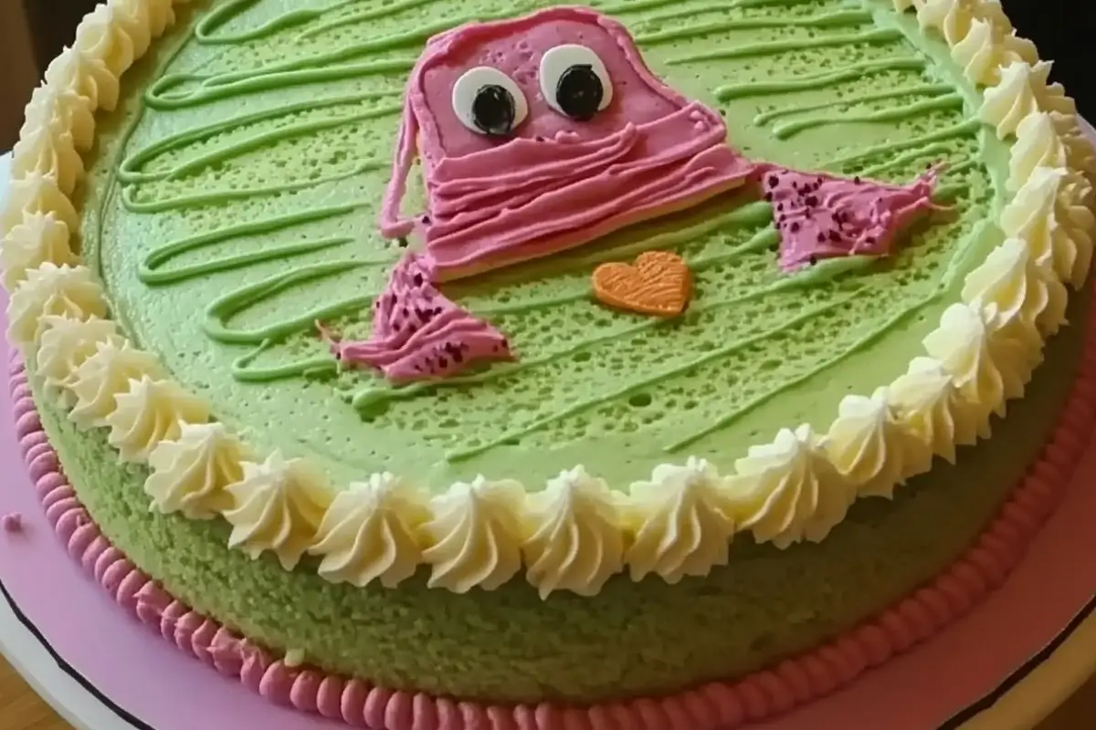 A colorful cake with green frosting, decorated with a whimsical pink frog-like character in the center, featuring large eyes and a small orange heart. The border of the cake has piped white frosting rosettes and a pink base trim.